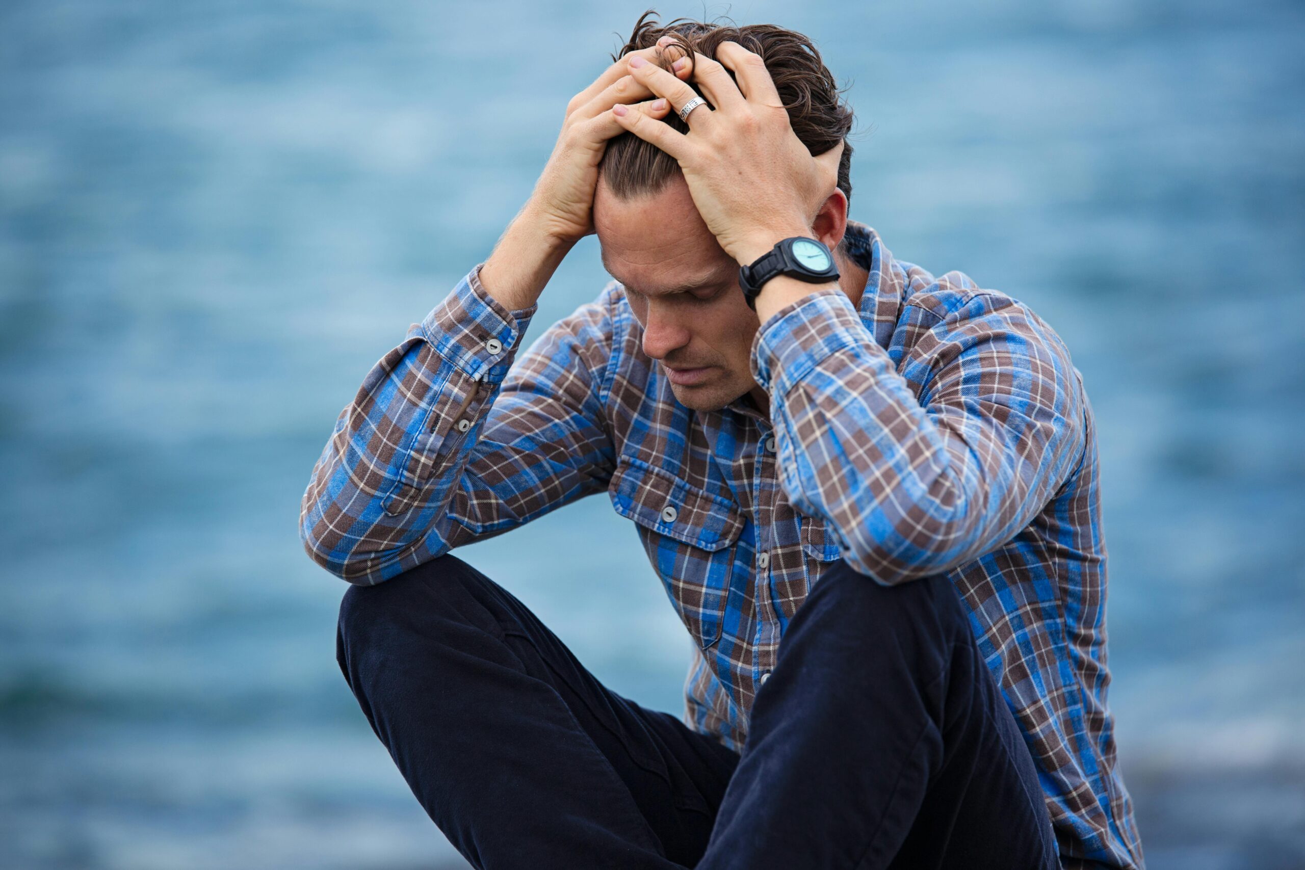 A man in a plaid shirt sits by the water looking distressed, symbolizing stress.